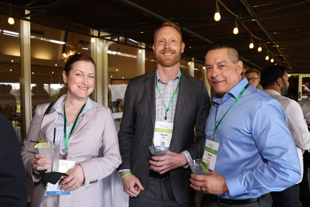 Three H2EA-NA conference attendees smiling and holding drinks while posing for a photo at a networking event; two men and one woman, all wearing name badges and green lanyards. This photo was taken at the North American Hydrogen Engine Conference.
