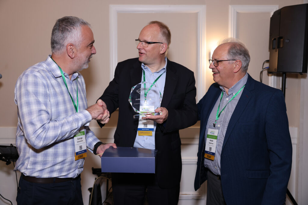 A speaker receives an award during the H2E-NA Conference banquet; three men with name badges and green lanyards are engaged in conversation, one handing over a crystal plaque. This photo was taken at the North American Hydrogen Engine Conference.